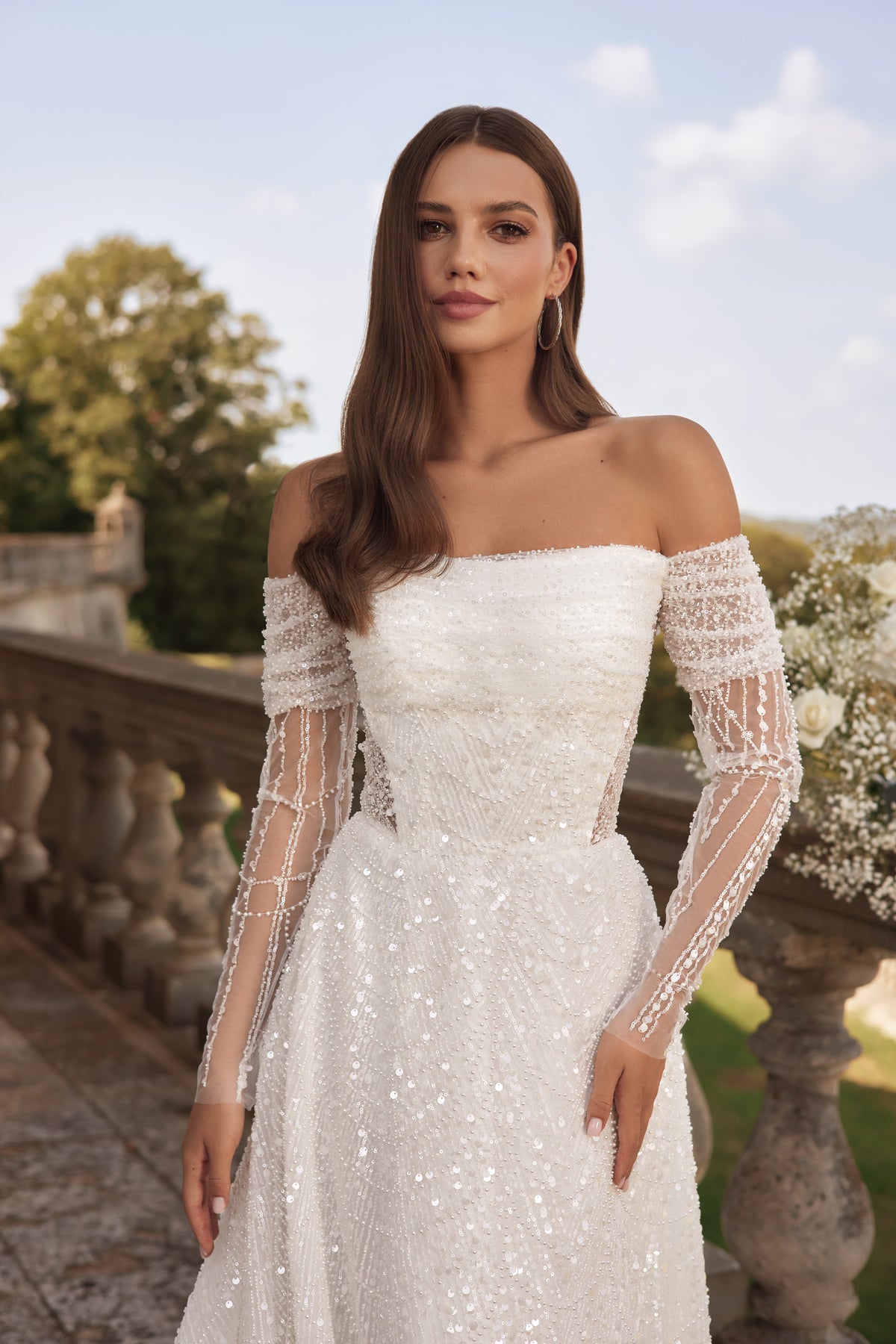 Close-up of an ivory wedding dress with a straight off-the-shoulder neckline, sheer beaded sleeves, and sparkle