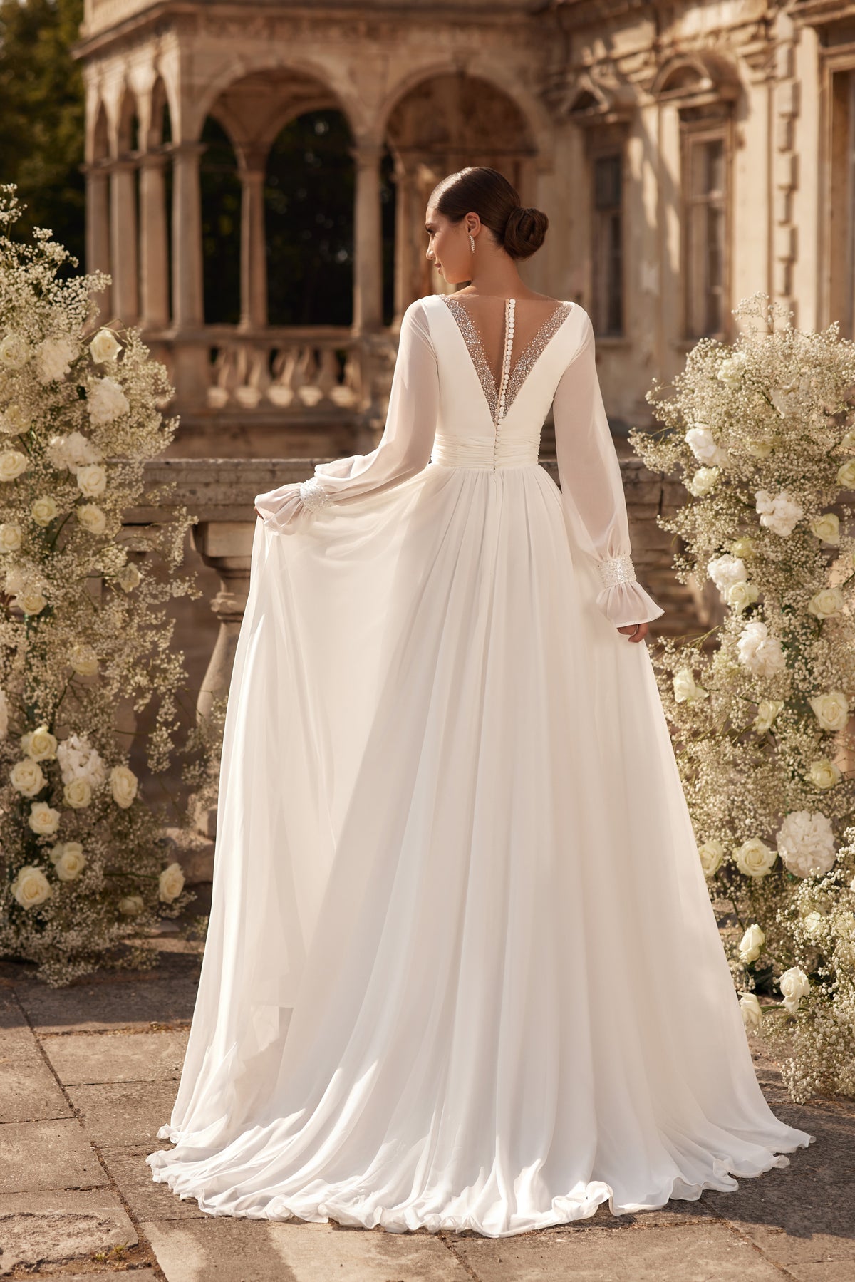 Ivory wedding dress showing a deep V-back with beaded edging, sheer sleeves, and a long flowing train