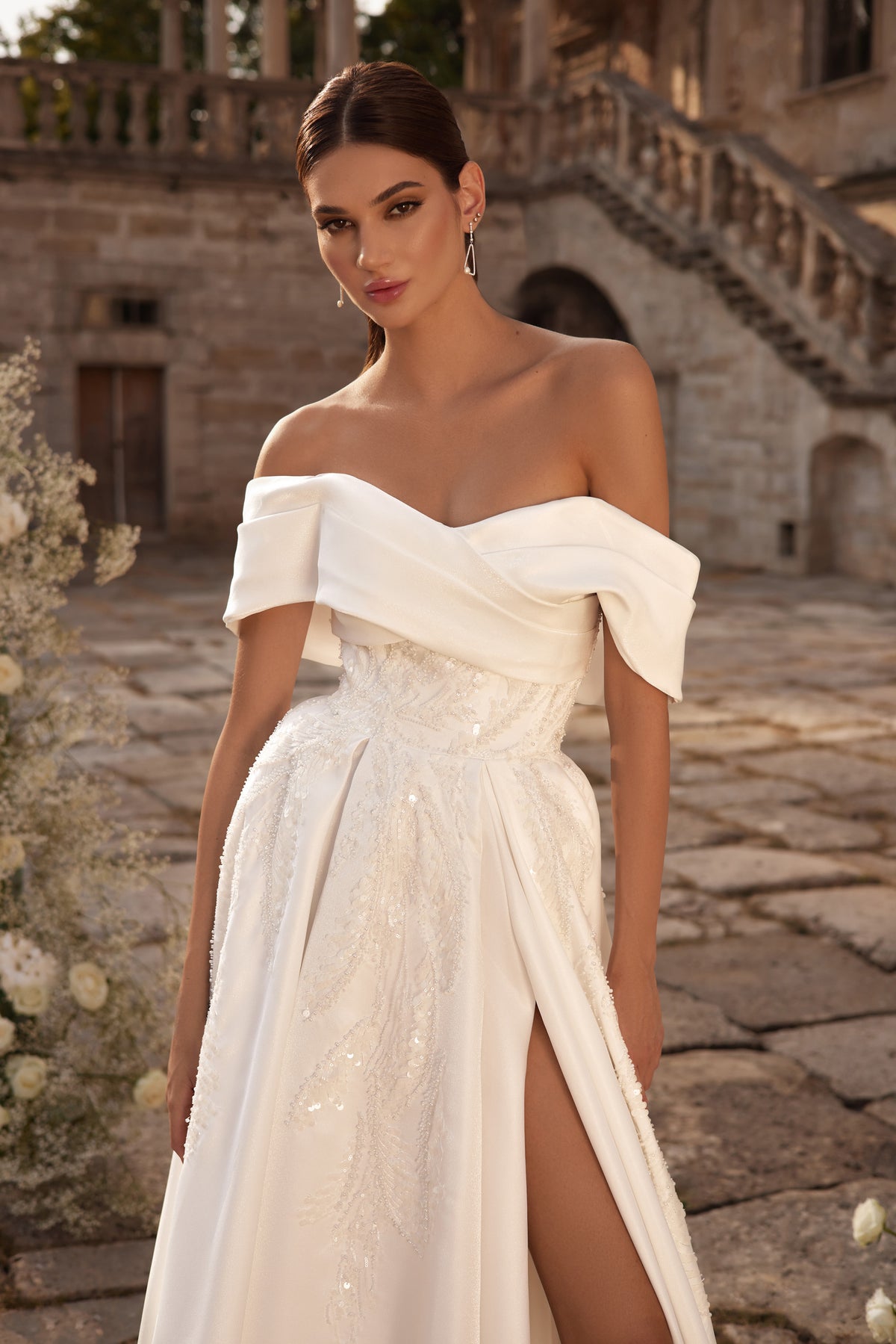 Close-up of an ivory wedding dress with a draped off-the-shoulder neckline, sparkling bodice, and thigh-high slit