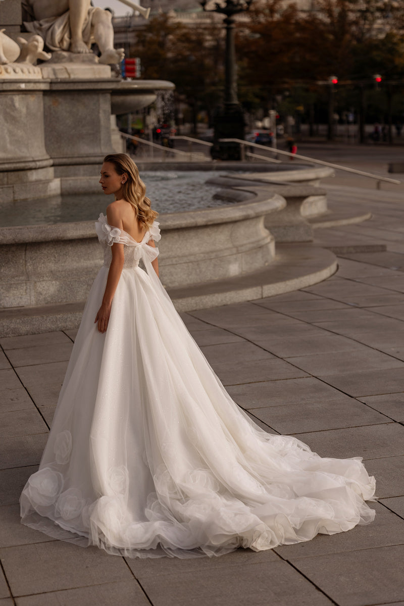 Ivory wedding dress showing an open back, off-the-shoulder floral sleeves, flowing A-line skirt, and train
