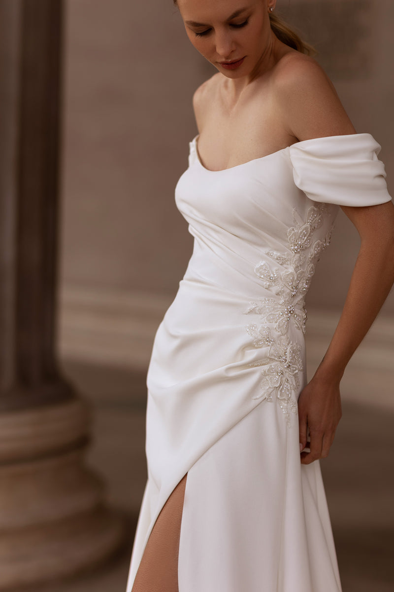 Close-up of an ivory wedding dress with ruched draping, floral appliqués, off-shoulder sleeves, and slit