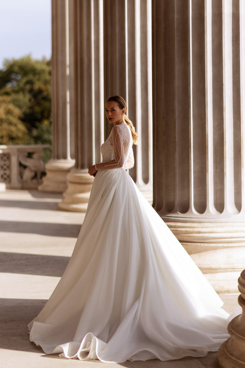 Three-quarter view of an ivory wedding dress with sheer beaded sleeves and a flowing full ball-gown skirt