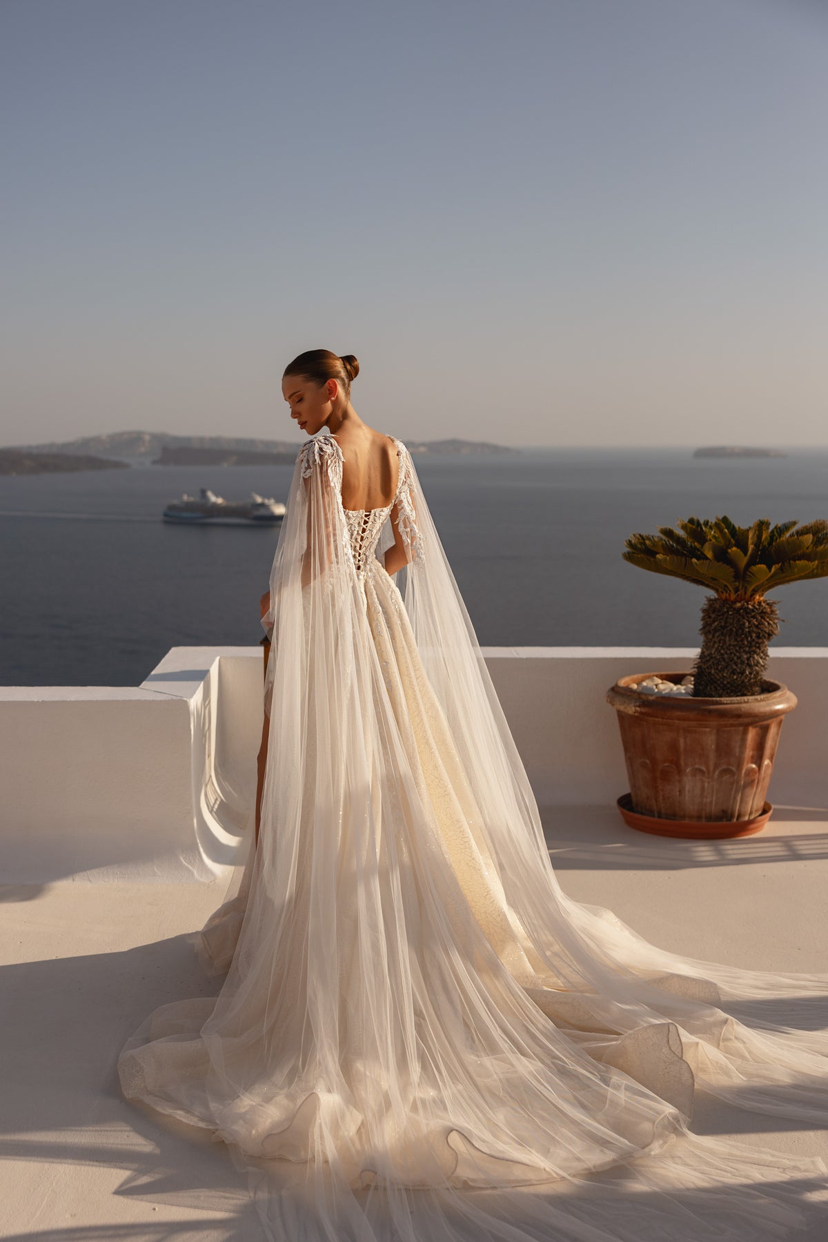 Open-back ivory wedding dress with a lace-up bodice, sheer cape, full skirt, and long trailing train