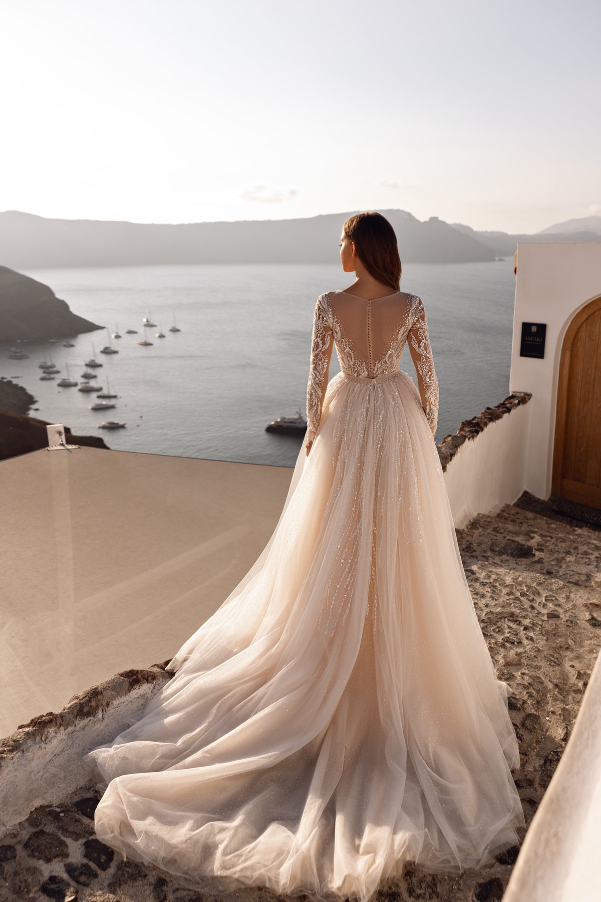 Back of an ivory wedding dress showing a sheer V-back, embellished sleeves, and a sweeping layered train