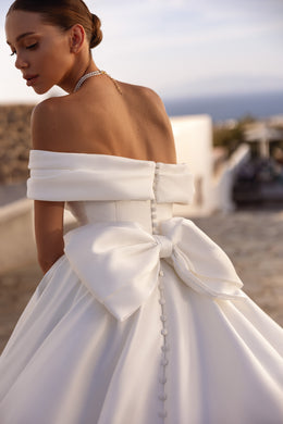 Open-back ivory wedding dress with an off-the-shoulder neckline, oversized bow, covered buttons, and a long train