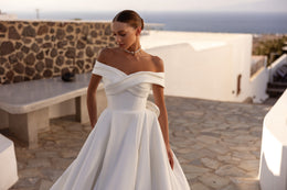 Ivory wedding dress featuring an off-the-shoulder crossover neckline, fitted waist, and softly pleated A-line skirt