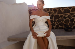 Seated ivory wedding dress with an off-the-shoulder crossover neckline and raised skirt revealing a front opening