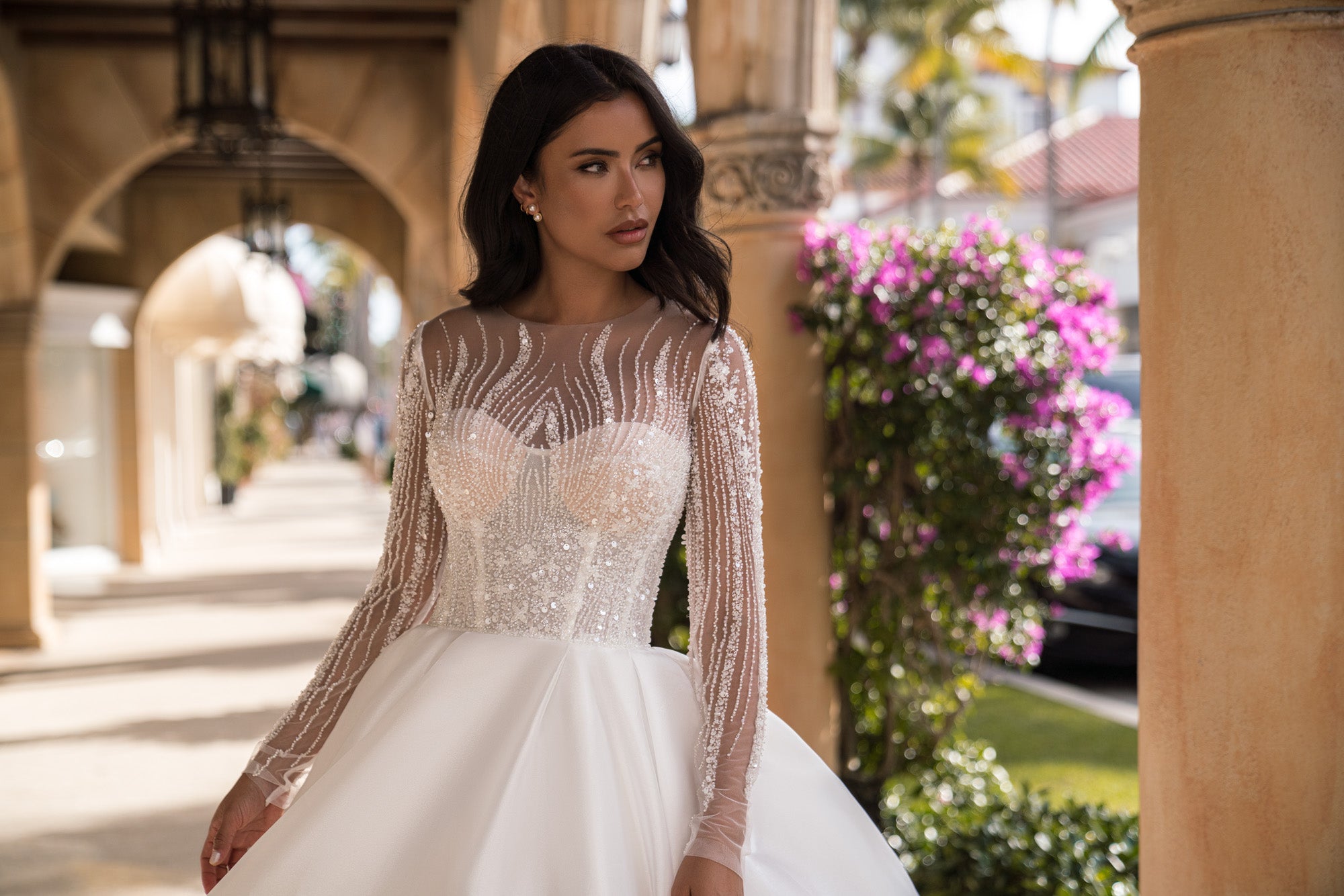 Close-up of a wedding dress bodice with a rounded neckline, sparkling beadwork, and sheer long sleeves