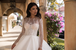Close-up of a wedding dress bodice with a rounded neckline, sparkling beadwork, and sheer long sleeves