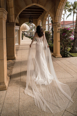 Back view of an ivory wedding dress with a sheer cape flowing into a long trailing train