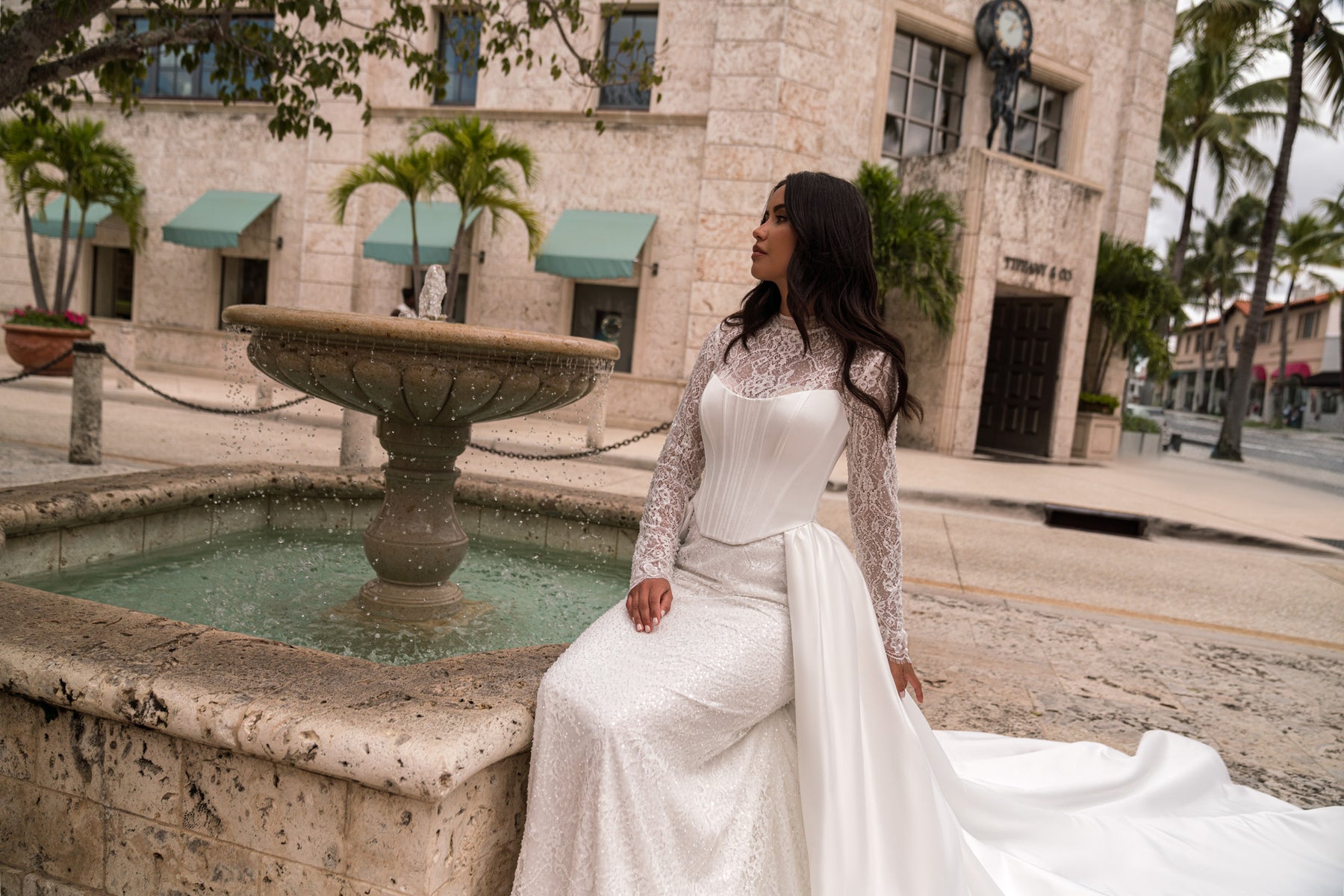 Ivory sheath wedding dress with a corset-style bodice, long lace sleeves, and draped overskirt