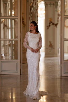 Fitted ivory wedding dress with a bateau neckline, glittering patterned fabric, sheer sleeves, and a narrow train