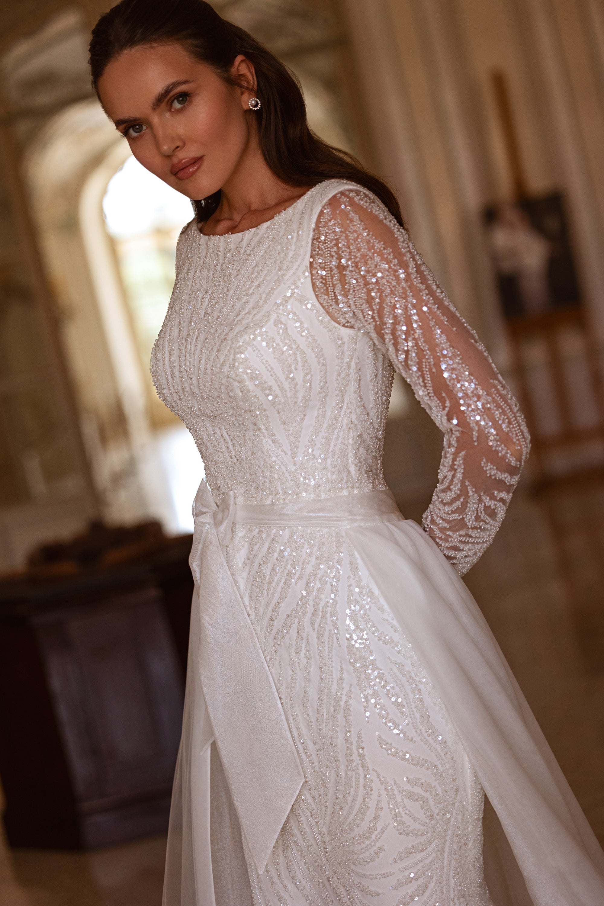 Close-up of swirling sequins on the bodice and sheer long sleeves of an ivory wedding dress with a draped overskirt