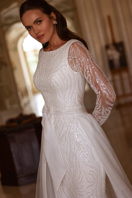 Close-up of swirling sequins on the bodice and sheer long sleeves of an ivory wedding dress with a draped overskirt