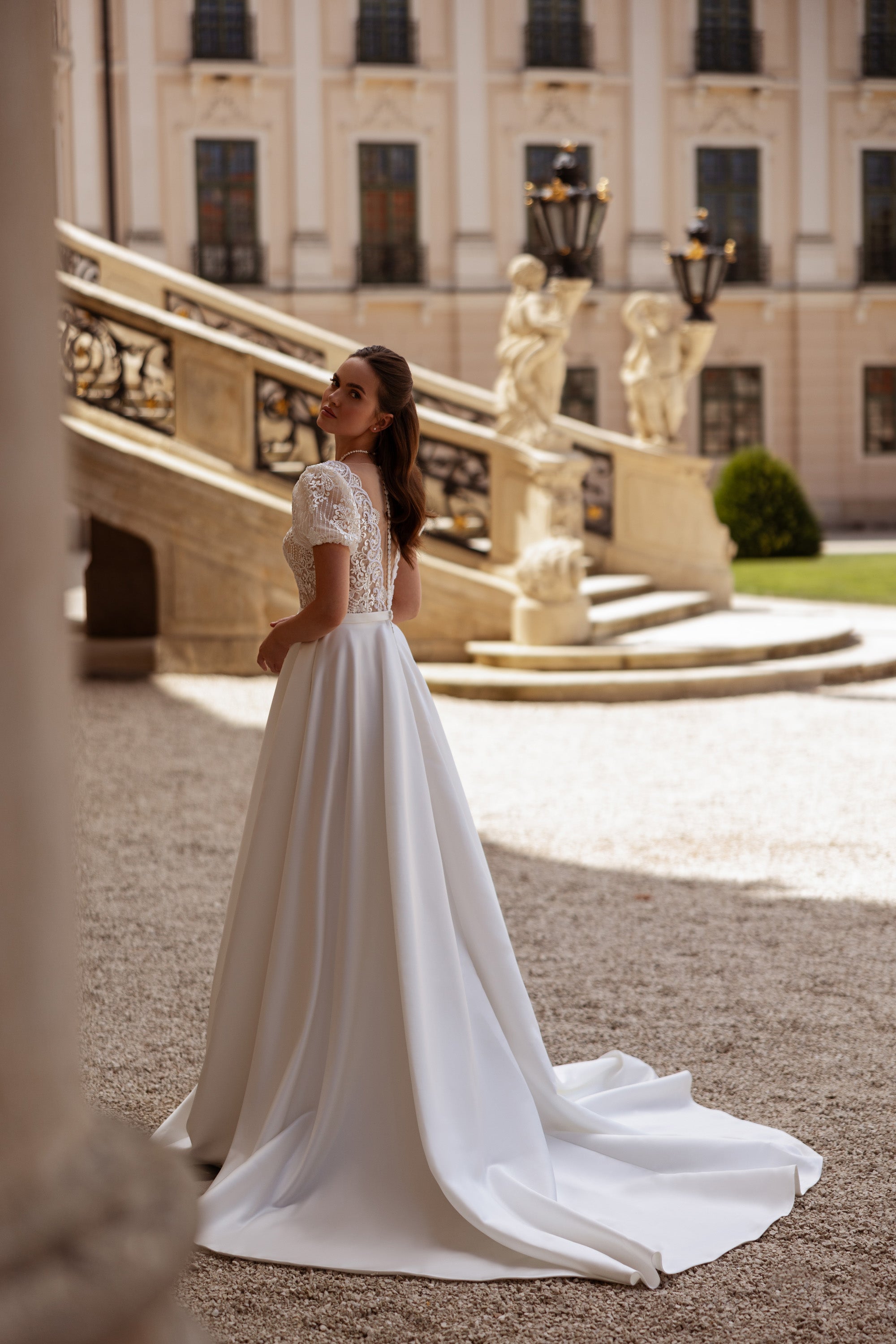 Rear three-quarter view of an ivory wedding dress with lace sleeves, open back, and trailing skirt