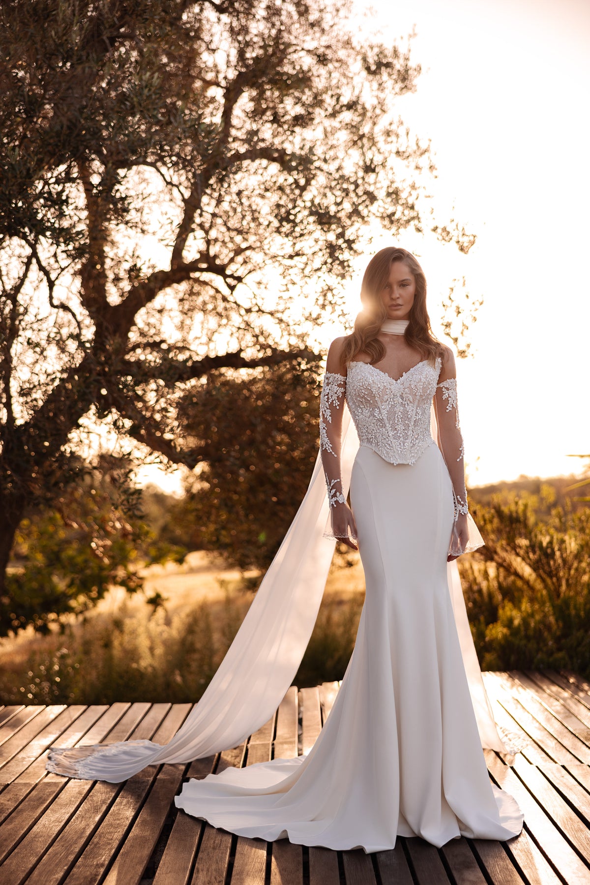 Full-length ivory wedding dress with an ornate lace bodice, sheer sleeves, fitted skirt, and sweeping cape