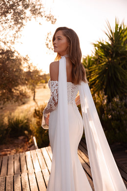 Side-back view of an ivory wedding dress with a fitted skirt, lace sleeves, and a long cape draped from the shoulders