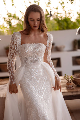 Ivory beaded sheath wedding dress with a square neckline, sheer long sleeves, and a tulle overskirt