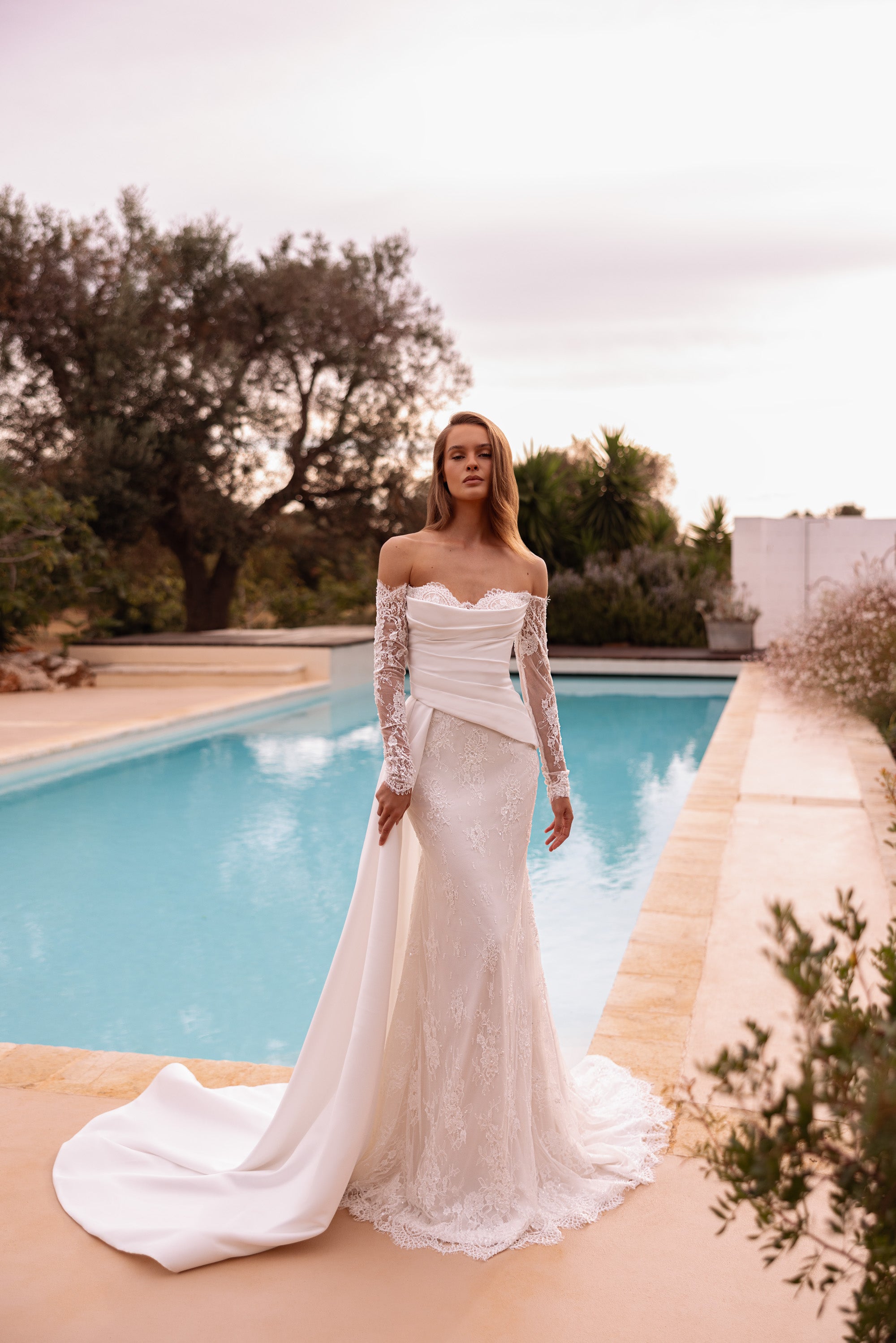 Ivory off-shoulder wedding dress with sheer lace sleeves, ruched bodice, mermaid skirt, and flowing overskirt