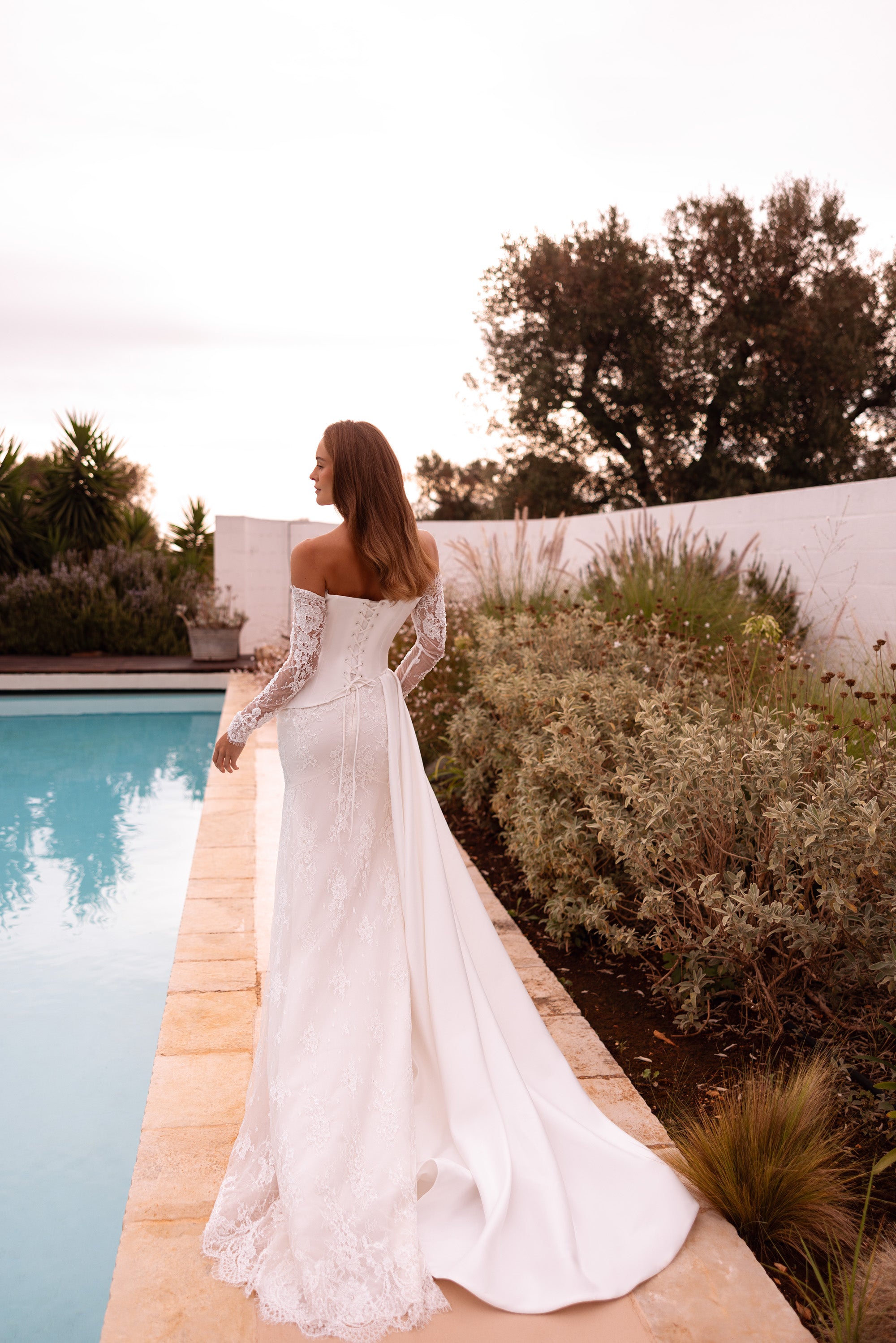 Back view of an ivory lace wedding dress with sheer sleeves, fitted mermaid skirt, and long draped overskirt