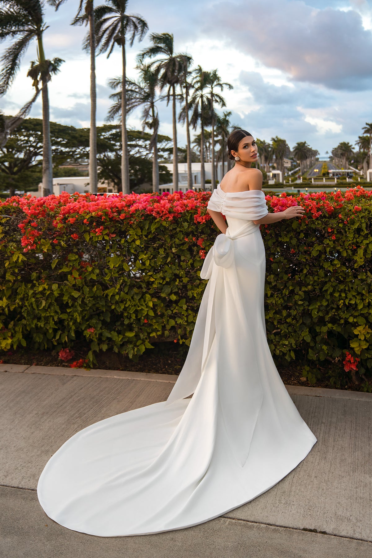 Ivory wedding dress shown from the back with a draped off-the-shoulder bolero, waist bow, fitted skirt, and sweeping train