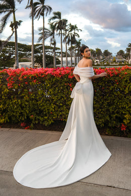 Ivory wedding dress shown from the back with a draped off-the-shoulder bolero, waist bow, fitted skirt, and sweeping train