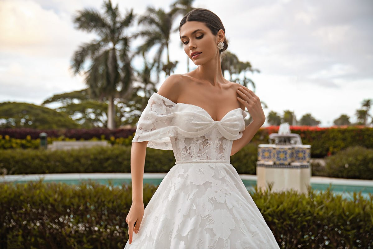 Close-up of a white wedding dress bodice with floral lace, off-the-shoulder sleeves, and a large bow