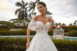 Close-up of a white wedding dress bodice with floral lace, off-the-shoulder sleeves, and a large bow