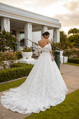 Back view of a white wedding dress with an off-the-shoulder neckline, waist bow, floral lace skirt, and long train