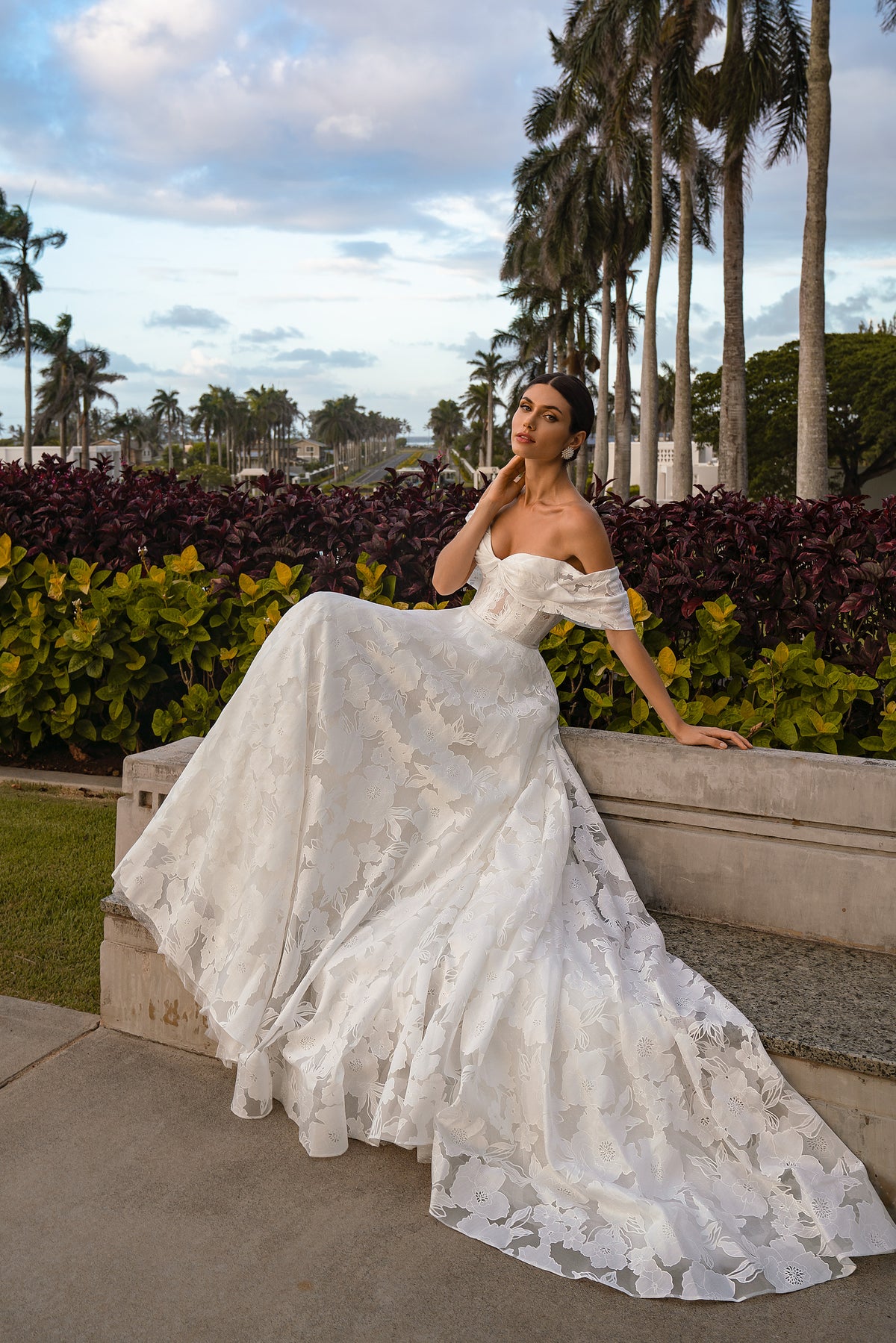 Seated white wedding dress with off-the-shoulder sleeves, floral lace ball gown skirt, and trailing hem