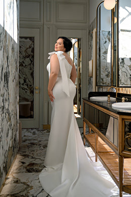 Ivory wedding dress with a large shoulder bow, fitted sheath silhouette, and sweeping back train