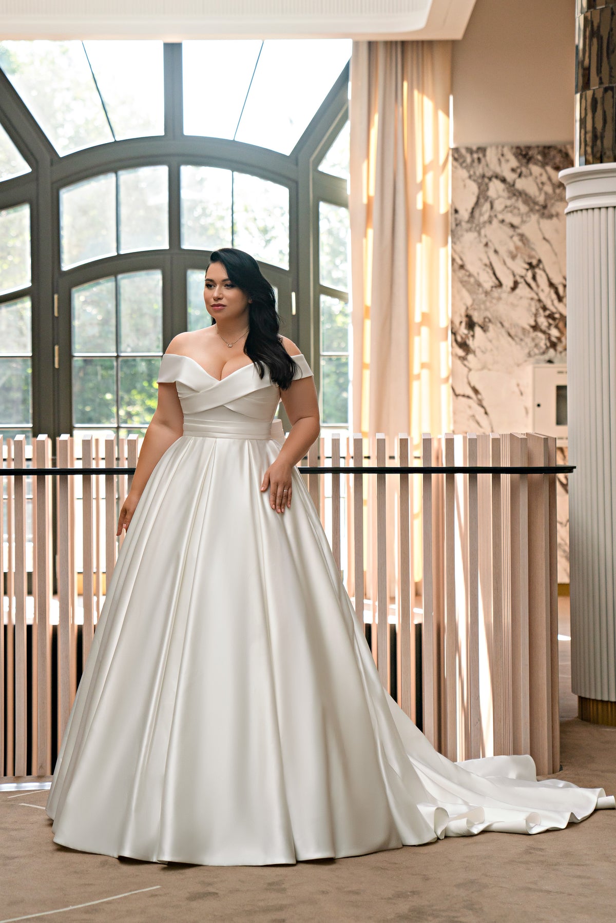 Off-the-shoulder ivory satin wedding dress with a wide pleated bodice, full skirt, and trailing train