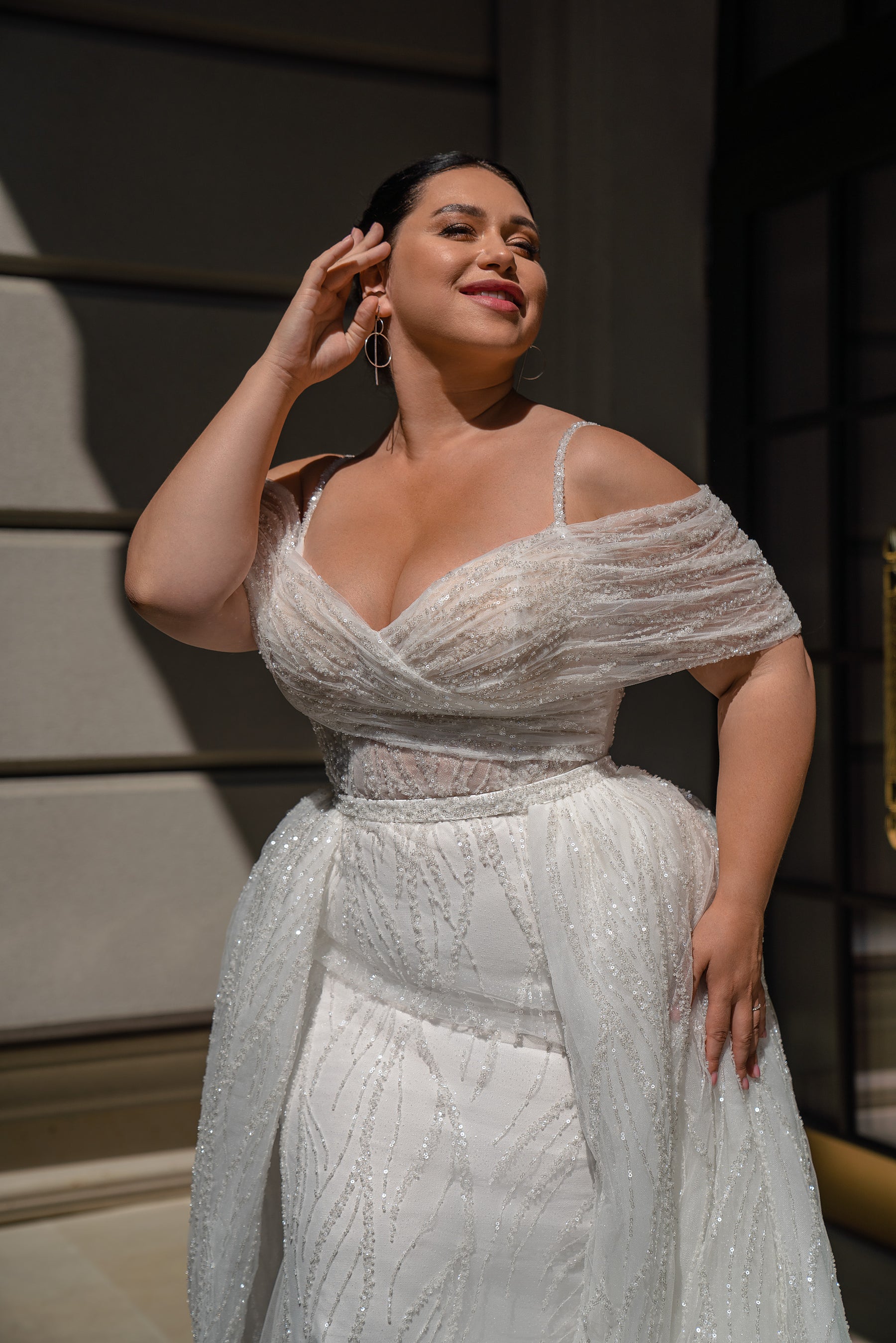 Ivory wedding dress with a draped off-shoulder V-neckline, thin straps, glittered bodice, and layered overskirt