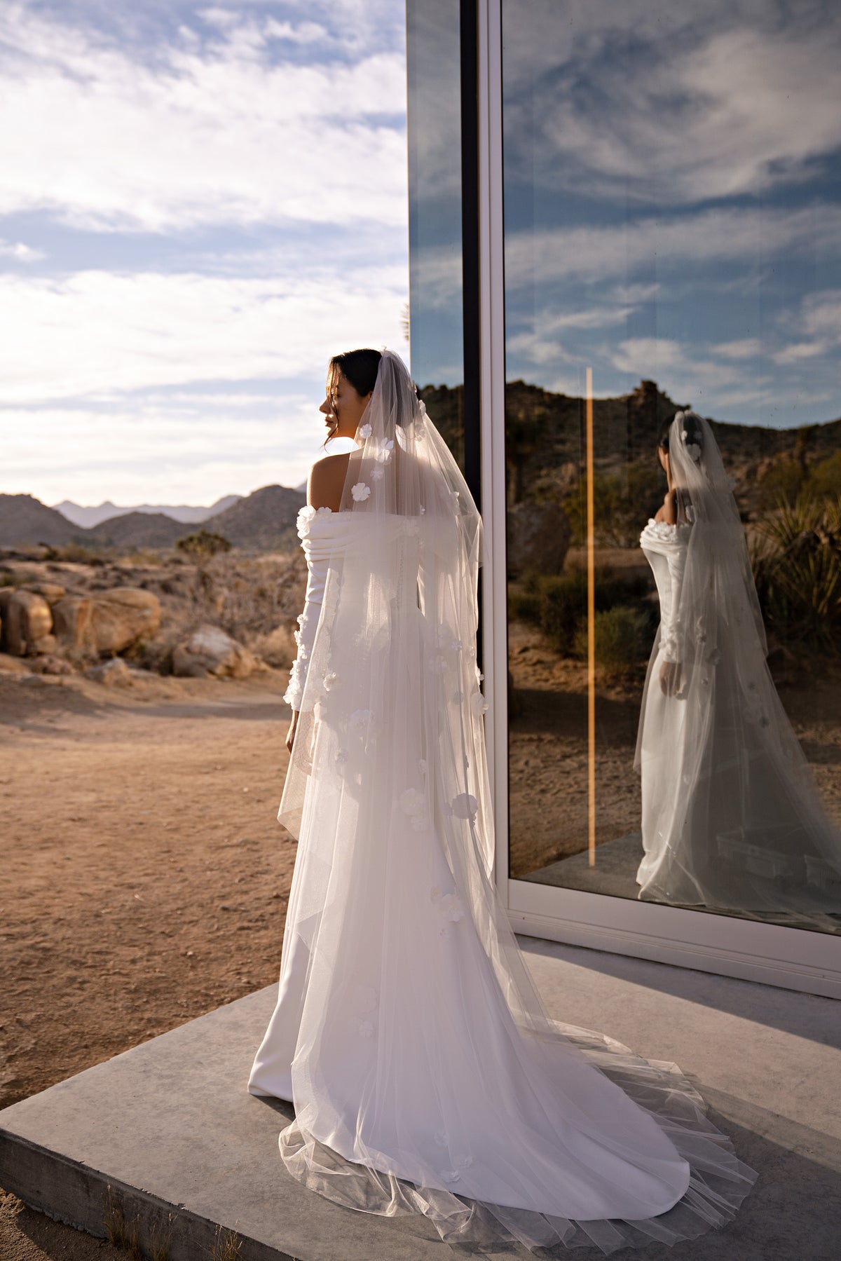 Side-profile ivory wedding dress with an off-the-shoulder neckline, long sleeves, fitted skirt, and trailing train