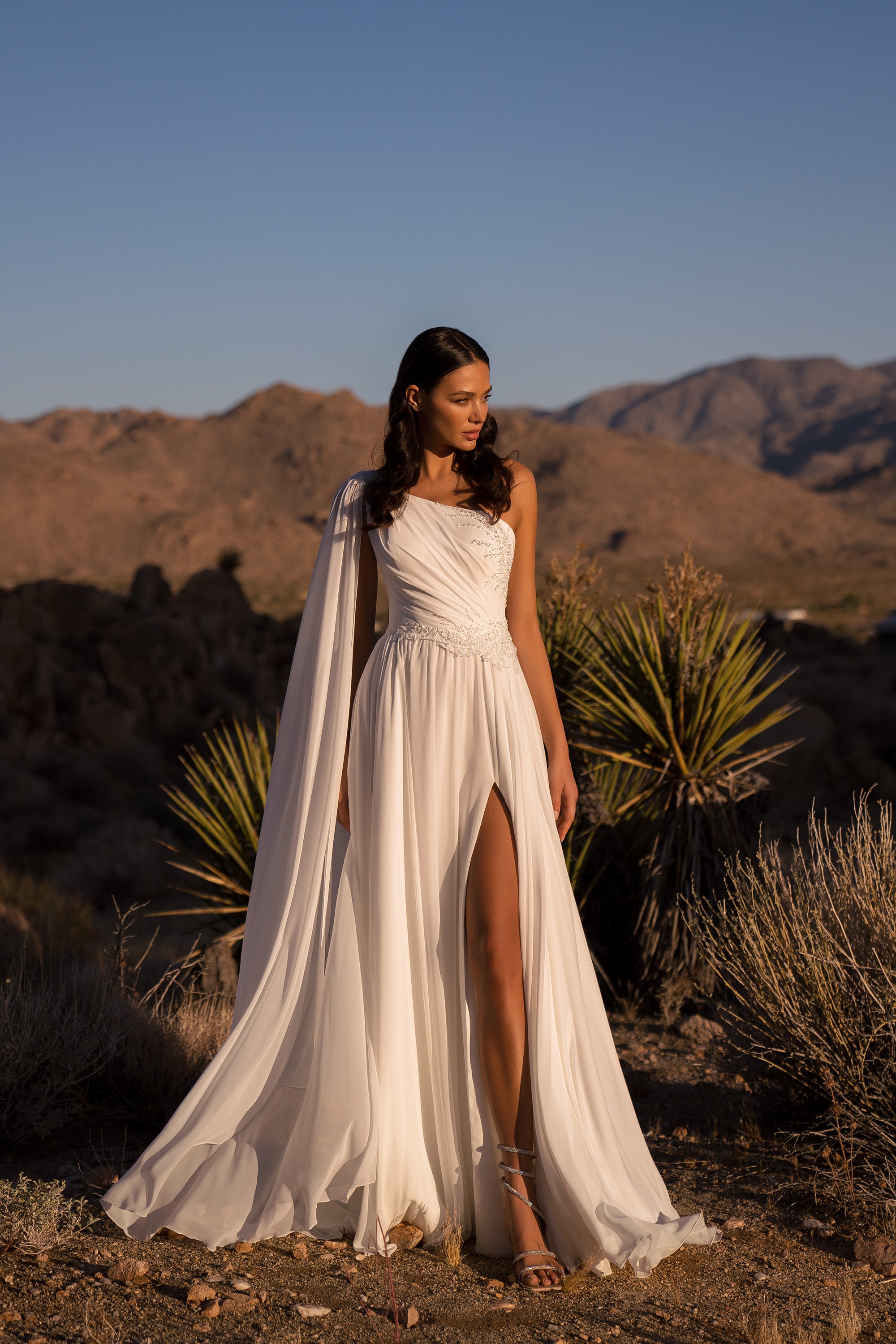 Ivory one-shoulder A-line wedding dress with a thigh-high slit and flowing draped cape