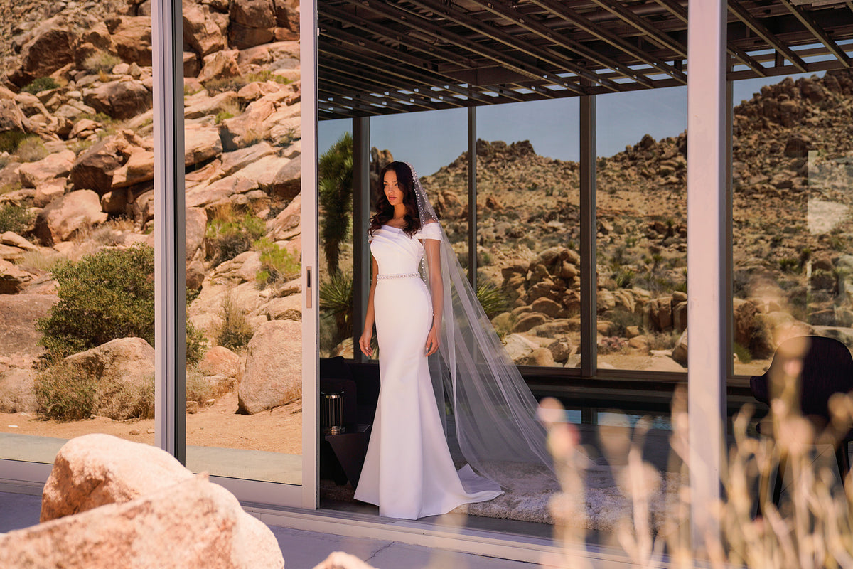 White wedding dress featuring a draped neckline, jeweled belt, fitted silhouette, and sweeping veil