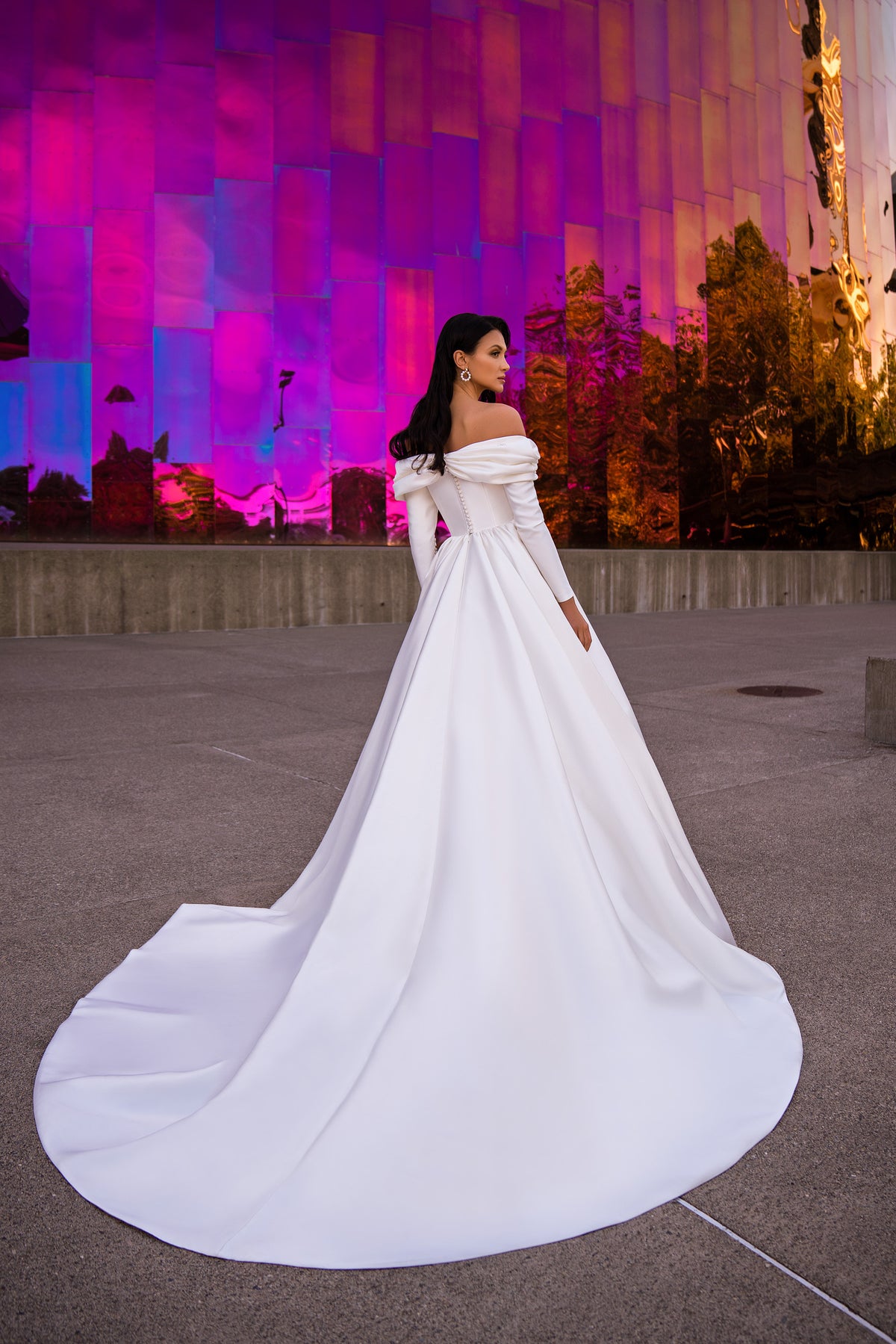 Three-quarter rear view of a white wedding dress with a sweeping train and draped off-the-shoulder neckline