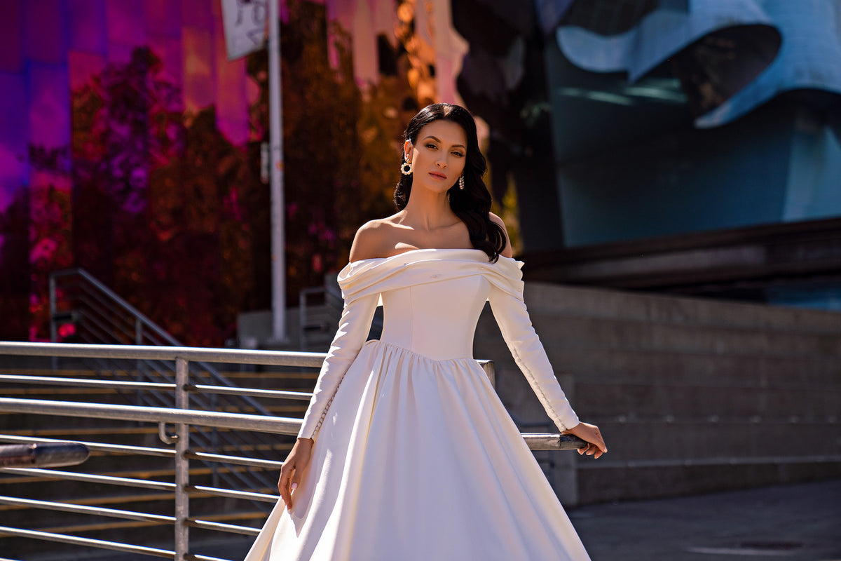 White wedding dress featuring a draped off-the-shoulder neckline, long sleeves, and a gathered waist