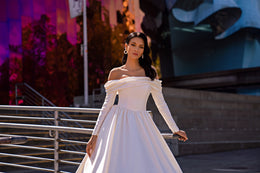 White wedding dress featuring a draped off-the-shoulder neckline, long sleeves, and a gathered waist