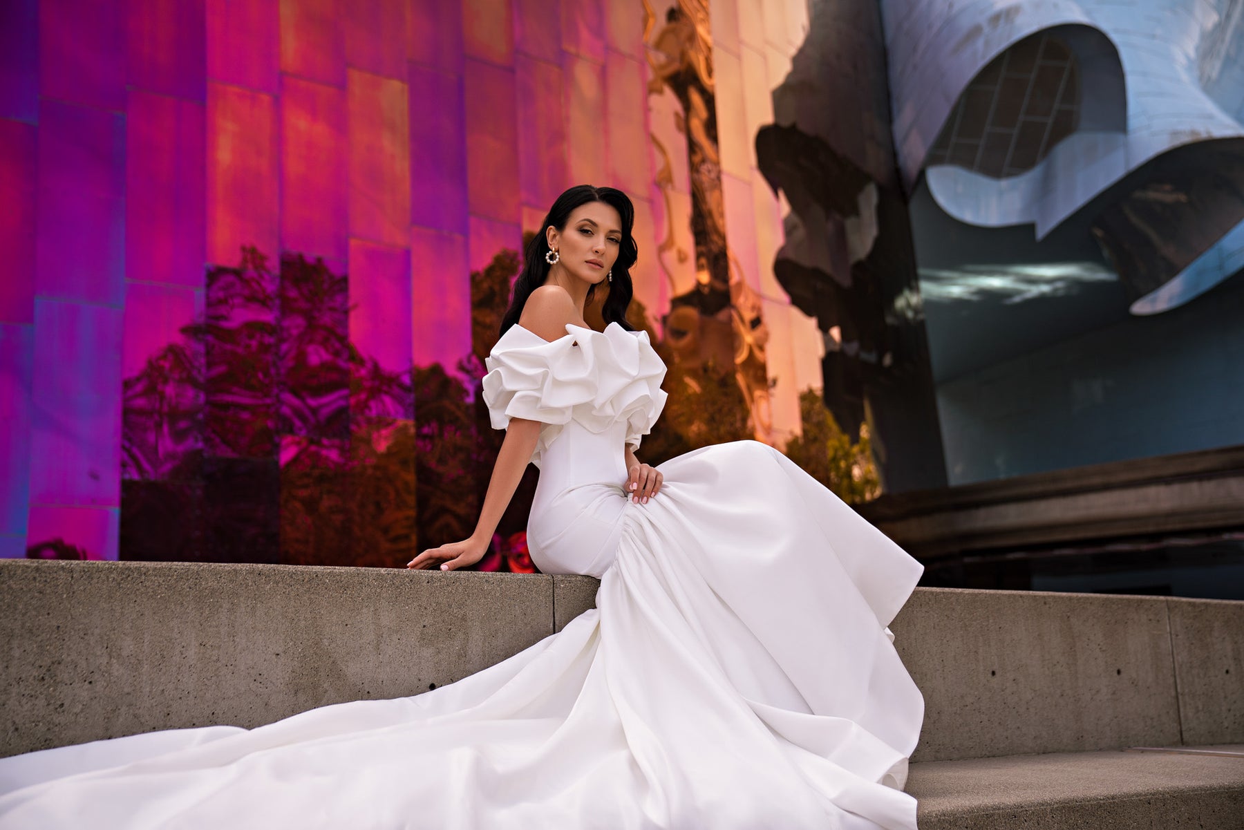 White wedding dress with a ruffled off-the-shoulder neckline, fitted bodice, and flowing skirt with train