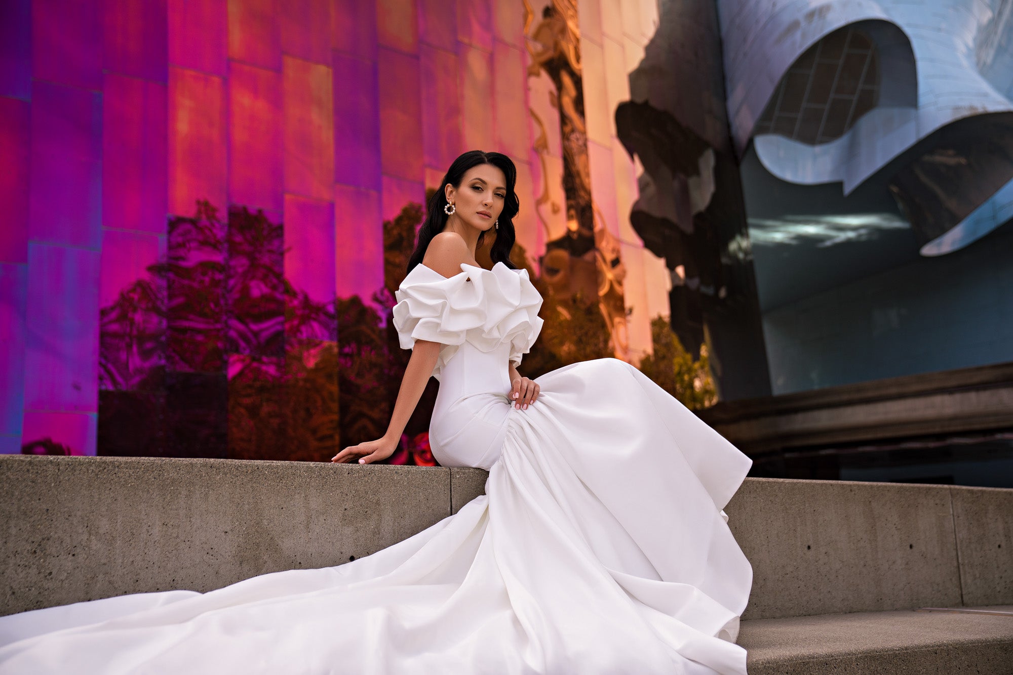 White wedding dress with a ruffled off-the-shoulder neckline, fitted bodice, and flowing skirt with train