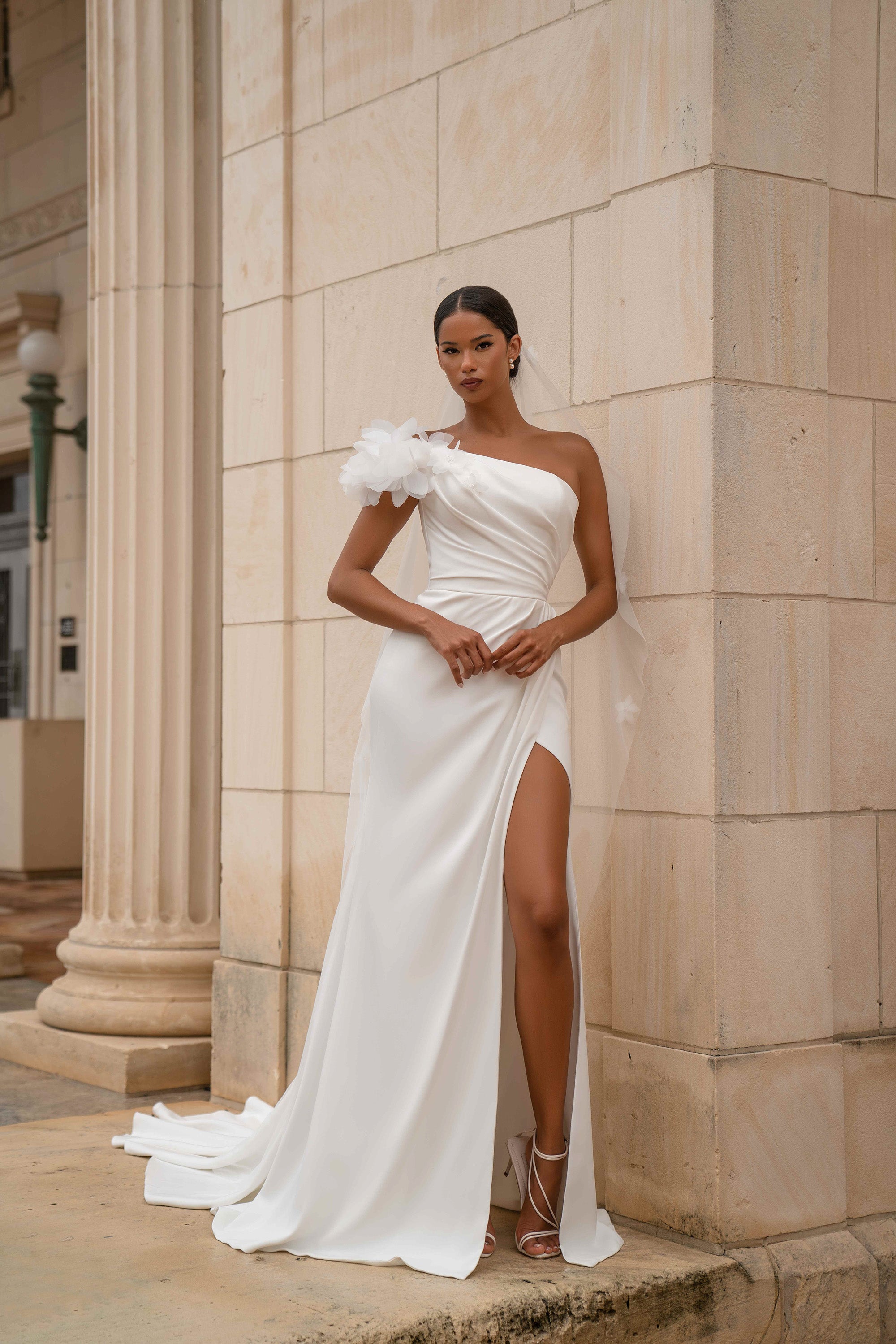 White one-shoulder wedding dress with a 3D floral shoulder, draped bodice, high slit, and train