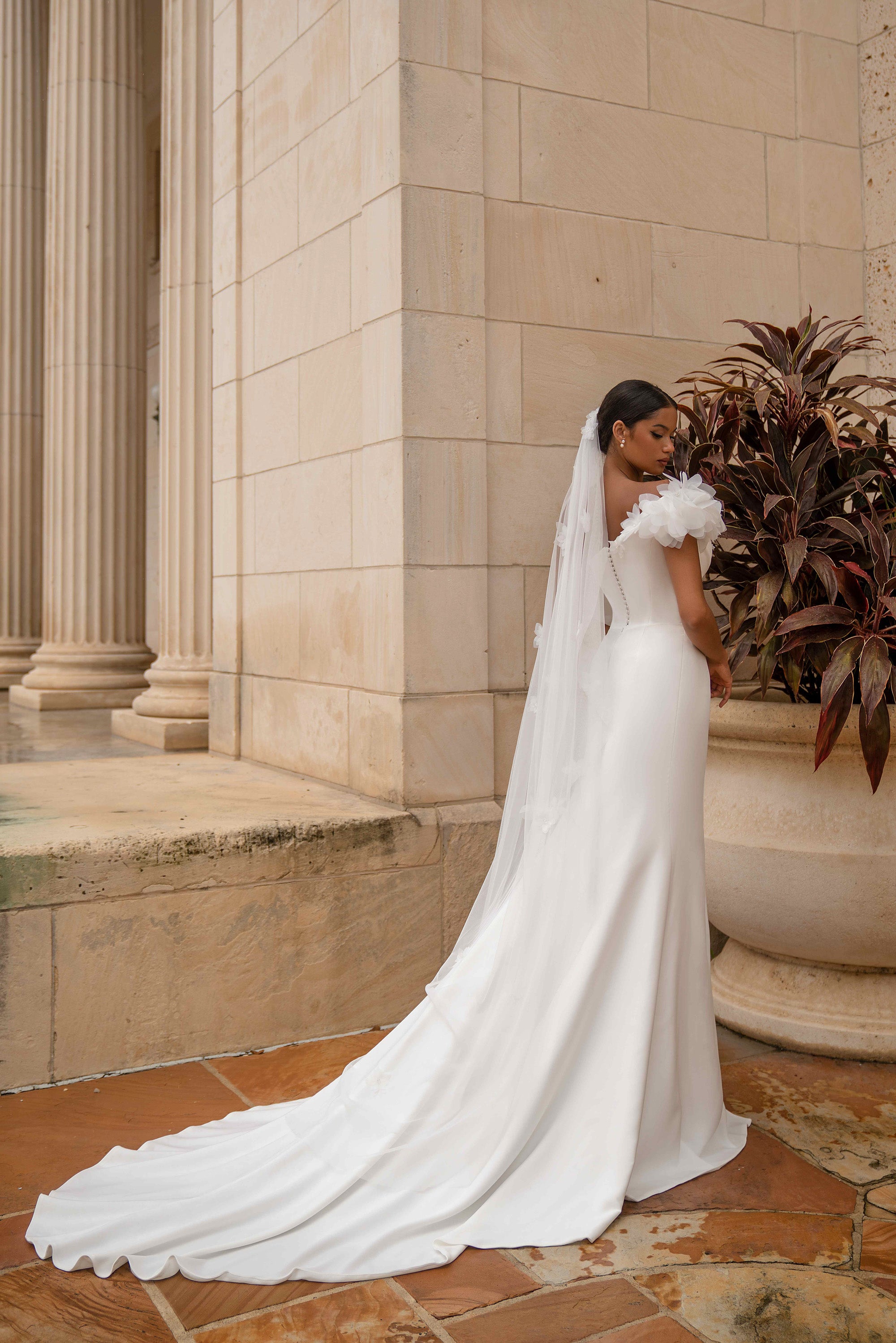 Rear view of a white one-shoulder wedding dress with a long flowing train and floral shoulder detail