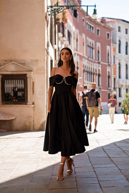 Black evening dress with an off-the-shoulder neckline, chain trim, thin straps, and a flowing pleated skirt