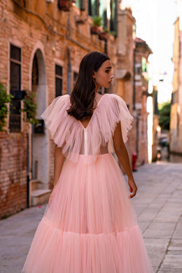Pink evening dress shown from the back with a V-shaped open back, sheer ruffled sleeves, and tiered skirt