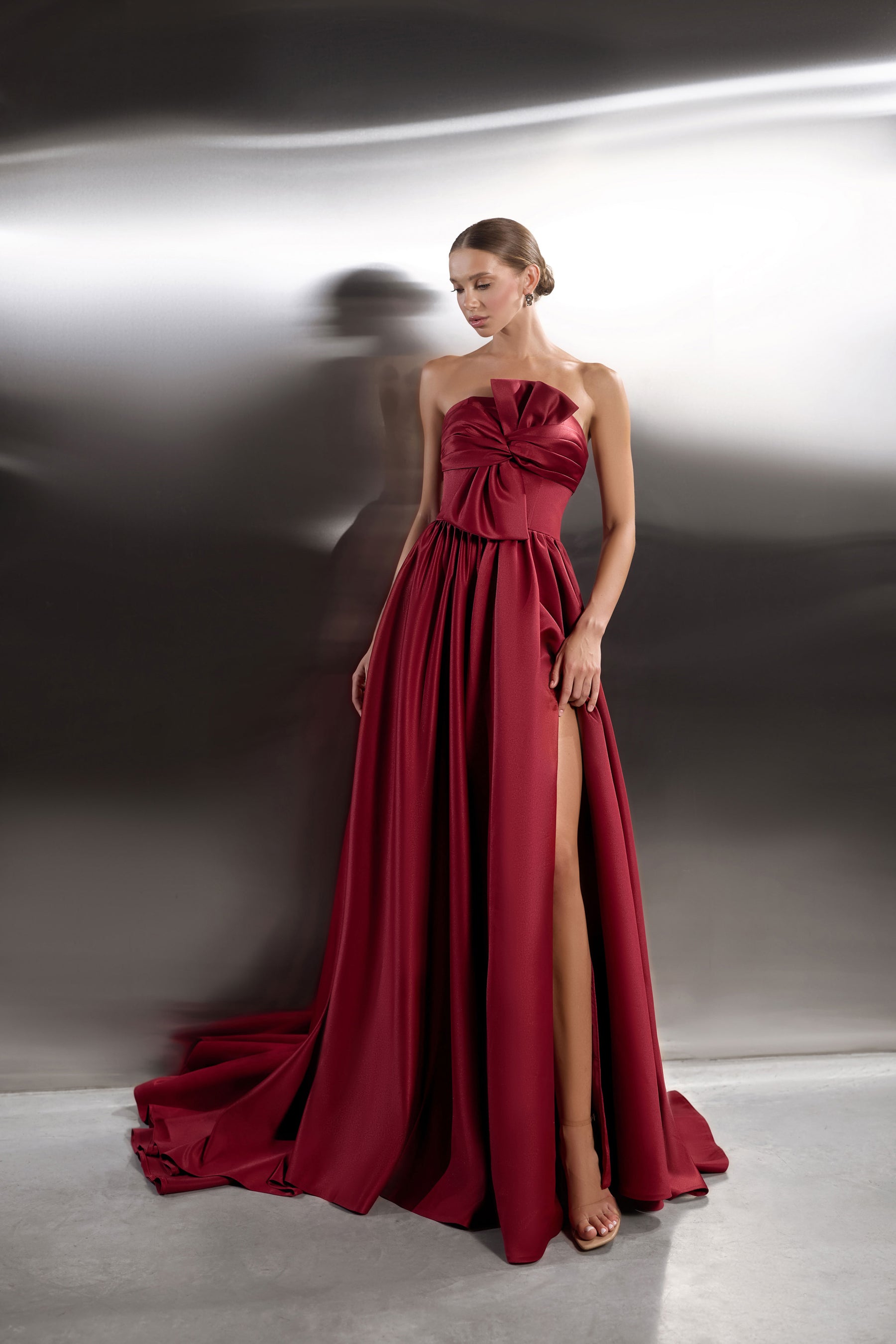 Burgundy strapless evening dress with a twisted bow bodice, thigh-high slit, full skirt, and flowing train