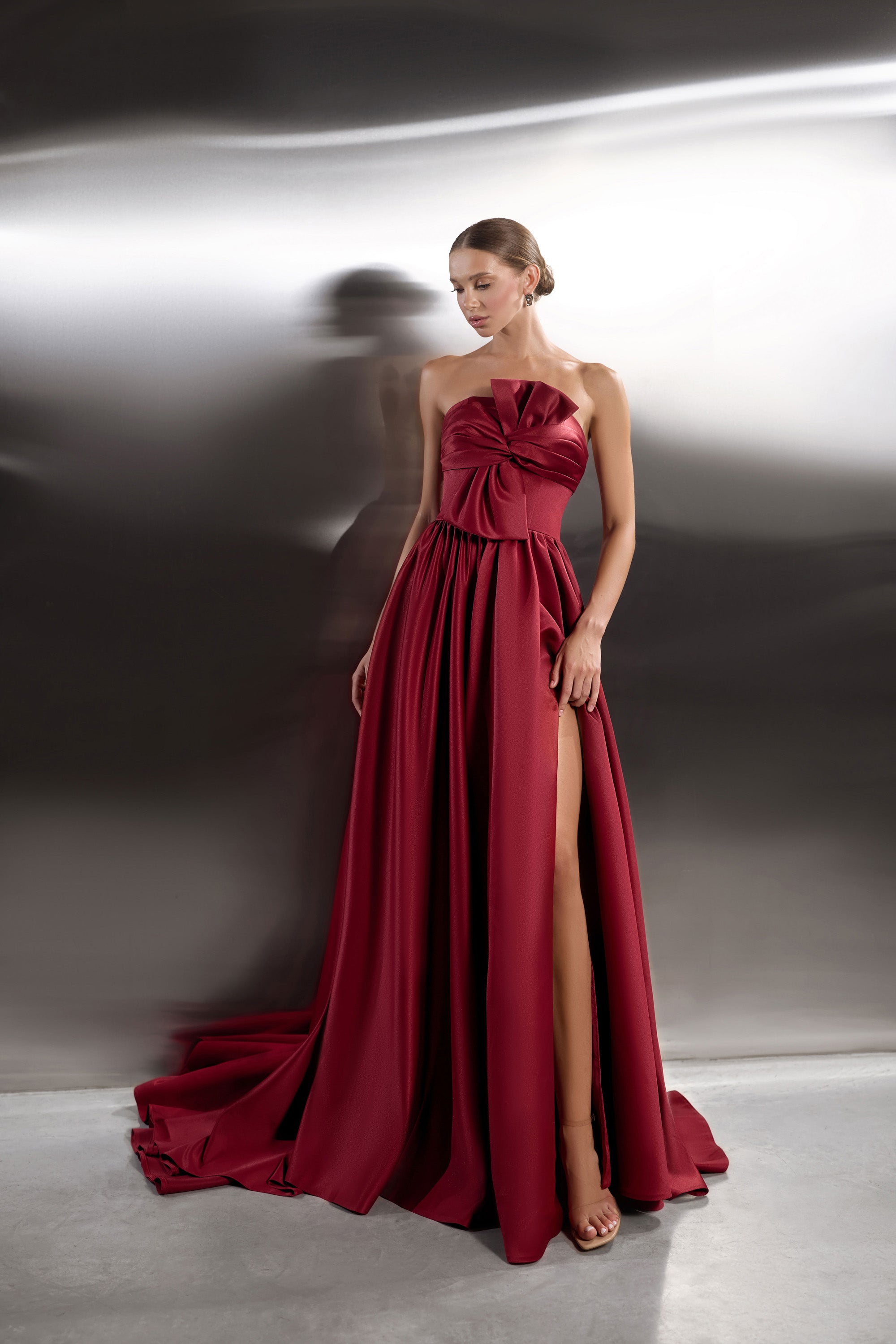 Burgundy strapless evening dress with a twisted bow bodice, thigh-high slit, full skirt, and flowing train