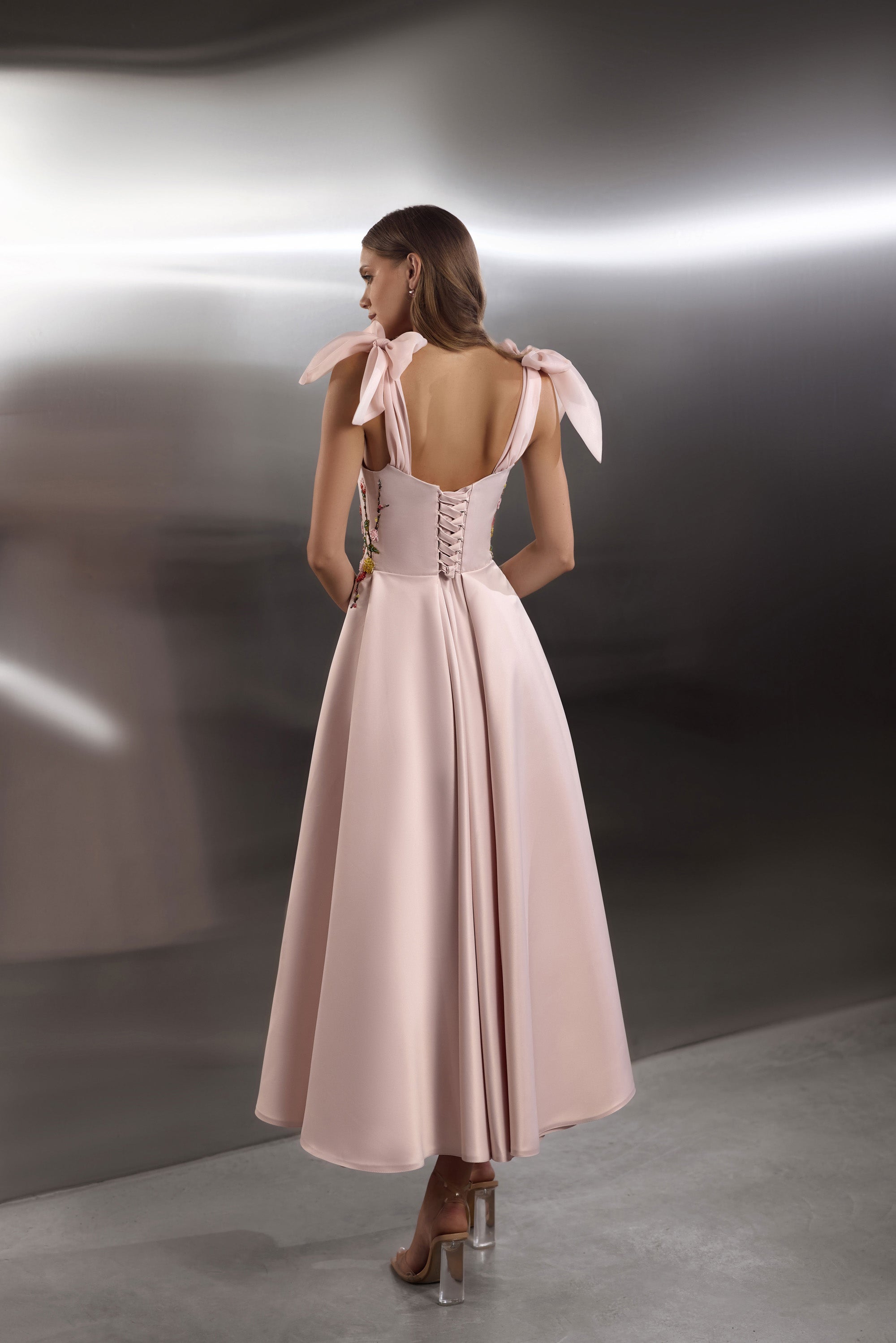 Blush pink evening dress showing an open back, corset-style lacing, ribbon shoulder ties, and a flowing skirt
