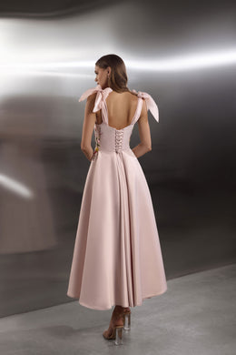 Blush pink evening dress showing an open back, corset-style lacing, ribbon shoulder ties, and a flowing skirt