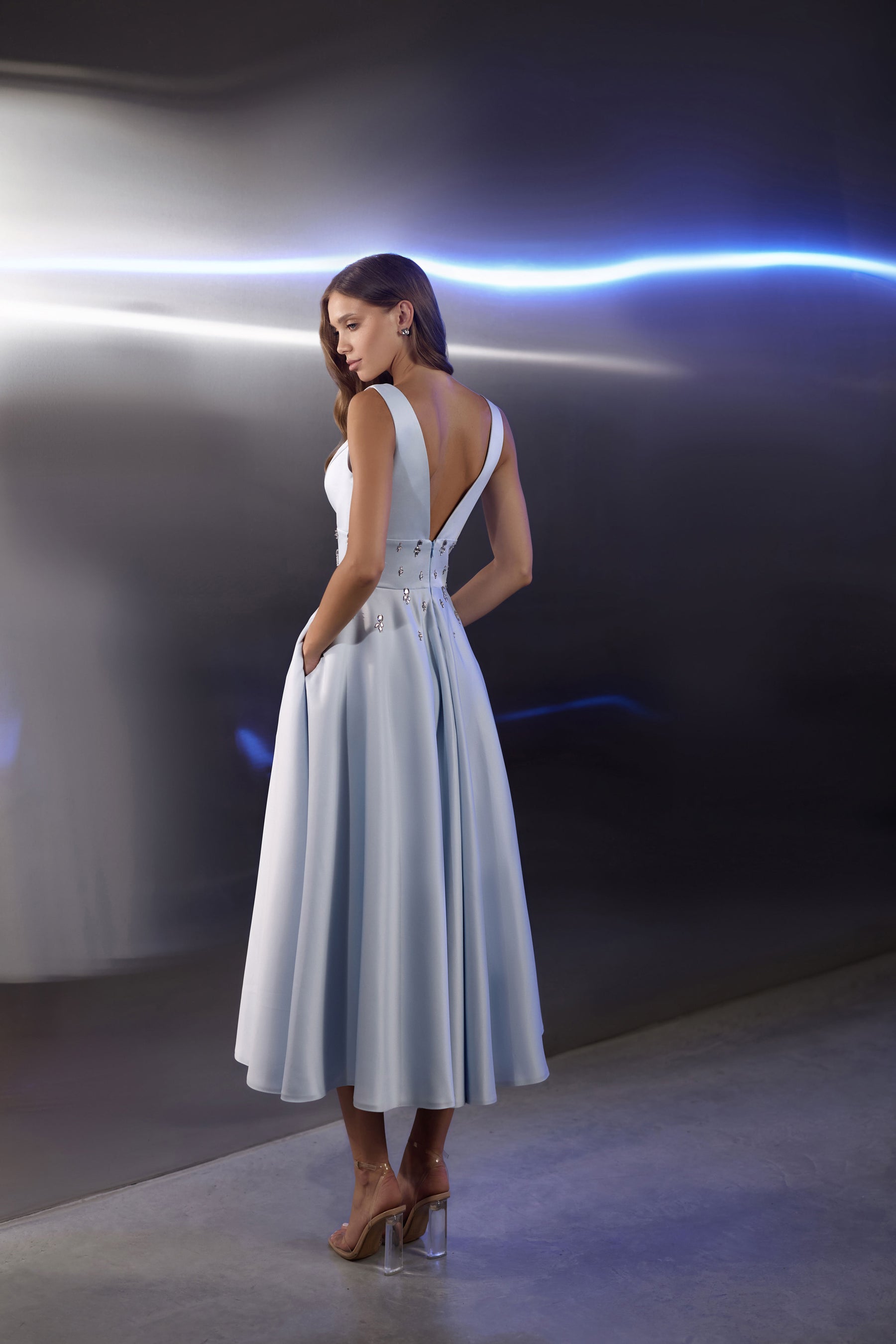 Open-back pale blue evening dress with crossing straps, jeweled waist details, and a flowing pleated skirt